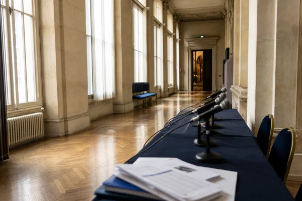 Couloir institutionnel parisien avec micros de presse et documents flous, dans une lumière naturelle claire.