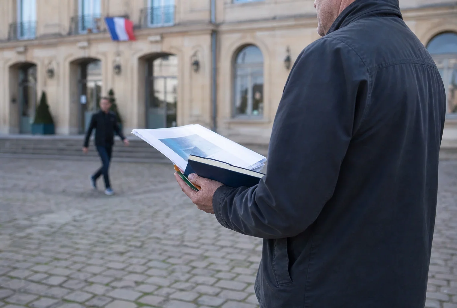 Un élu local de dos tient un dossier devant la mairie de Saint-Avold, en lumière naturelle claire.