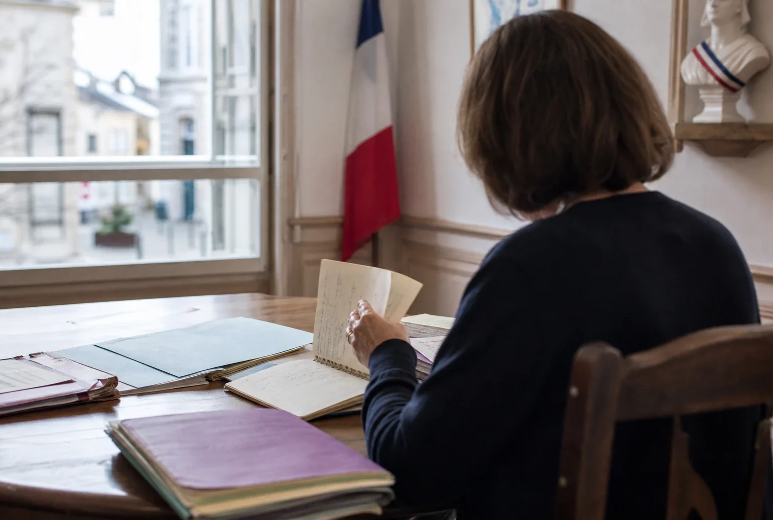 Élu local anonyme de dos dans une mairie, carnet et dossier en main, lumière naturelle de fin de journée.