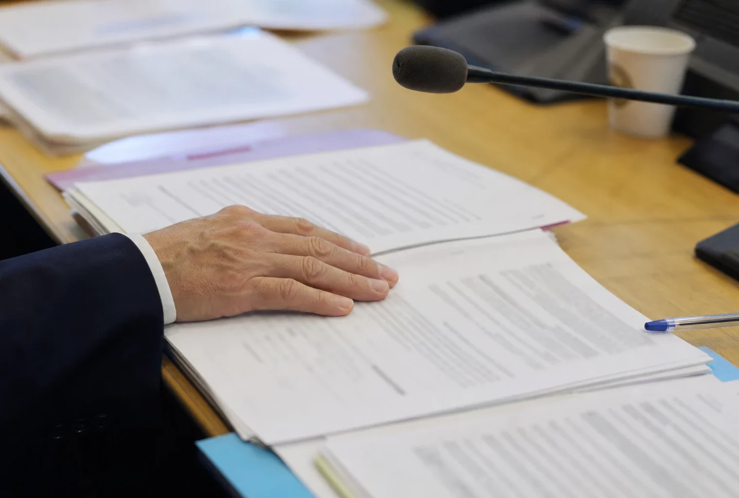 Main anonyme, micro de commission et dossier flou sur une table de salle d’audition parlementaire éclairée par une fenêtre.