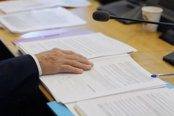 Main anonyme, micro de commission et dossier flou sur une table de salle d’audition parlementaire éclairée par une fenêtre.