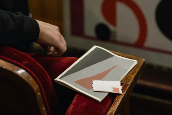 Main anonyme sur un fauteuil de salle, programme culturel flou et billet sans texte lisible dans un théâtre français.