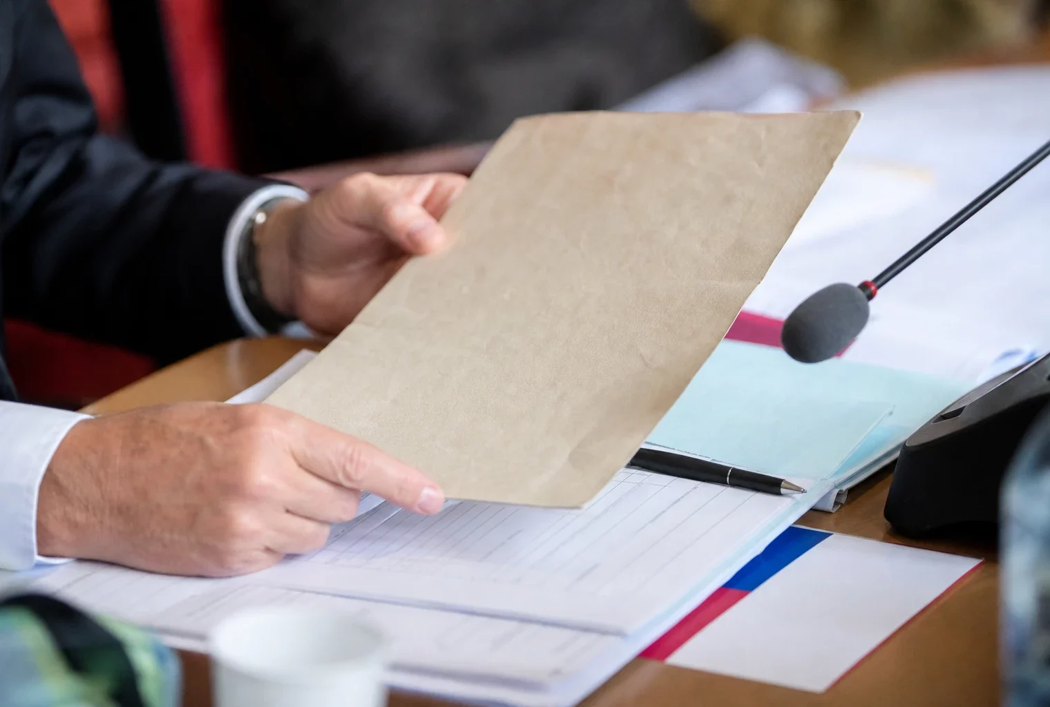 Dossier de commission parlementaire tenu par des mains anonymes, avec micro et documents flous sous lumière naturelle.