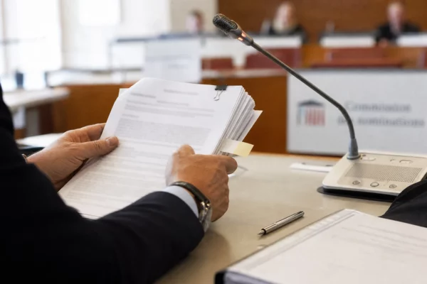 Dossier de commission tenu à la main près d’un micro, dans une salle institutionnelle française lumineuse.