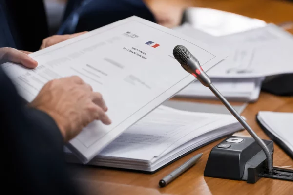 Mains anonymes feuilletant un dossier lors d’une audition, avec micro de commission et documents flous en premier plan.