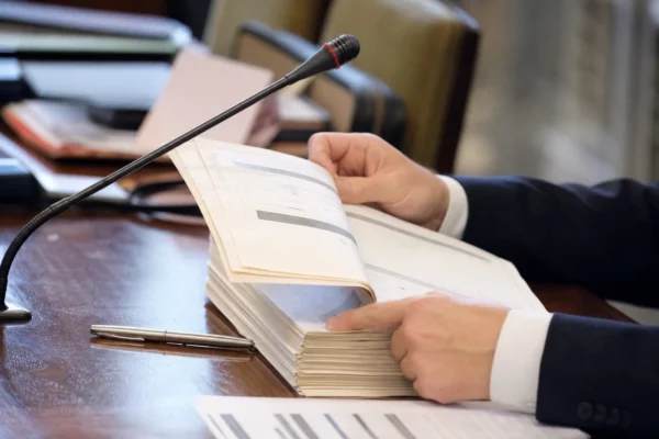 Dossier d’audition tenu par des mains anonymes avec micro de commission dans une salle parlementaire floue