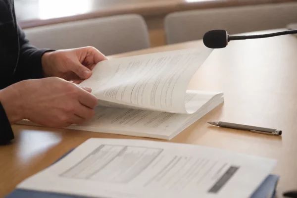 Mains anonymes feuilletant un dossier de nomination devant un micro de commission parlementaire