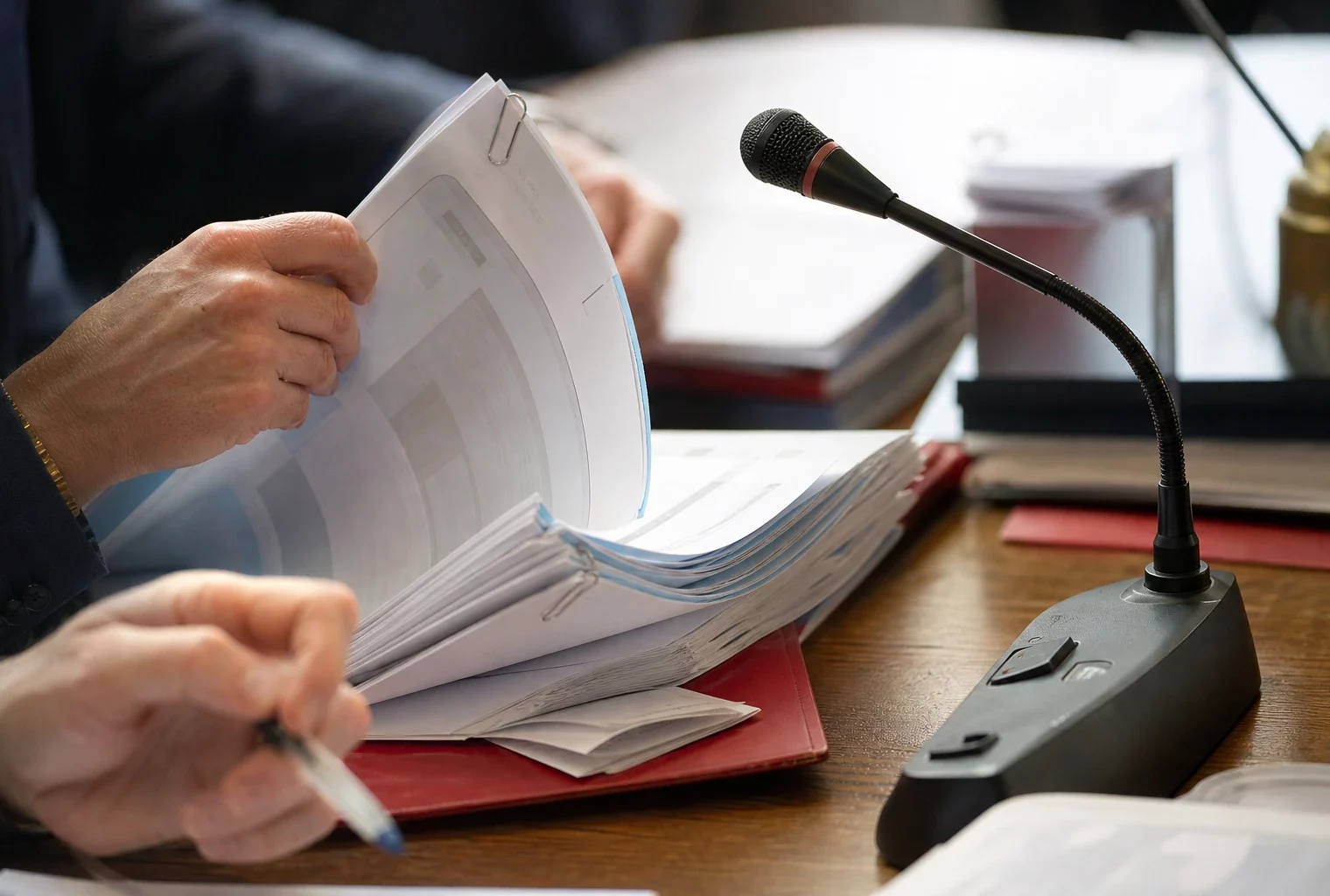 Mains anonymes feuilletant un dossier en commission parlementaire, avec micro de table et documents flous.