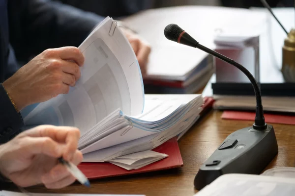 Mains anonymes feuilletant un dossier en commission parlementaire, avec micro de table et documents flous.