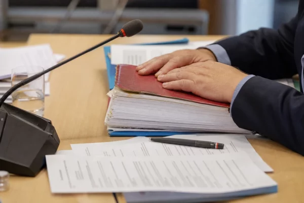 Dossier et micro de commission parlementaire sur une table, avec des mains anonymes et des documents flous en lumière naturelle.