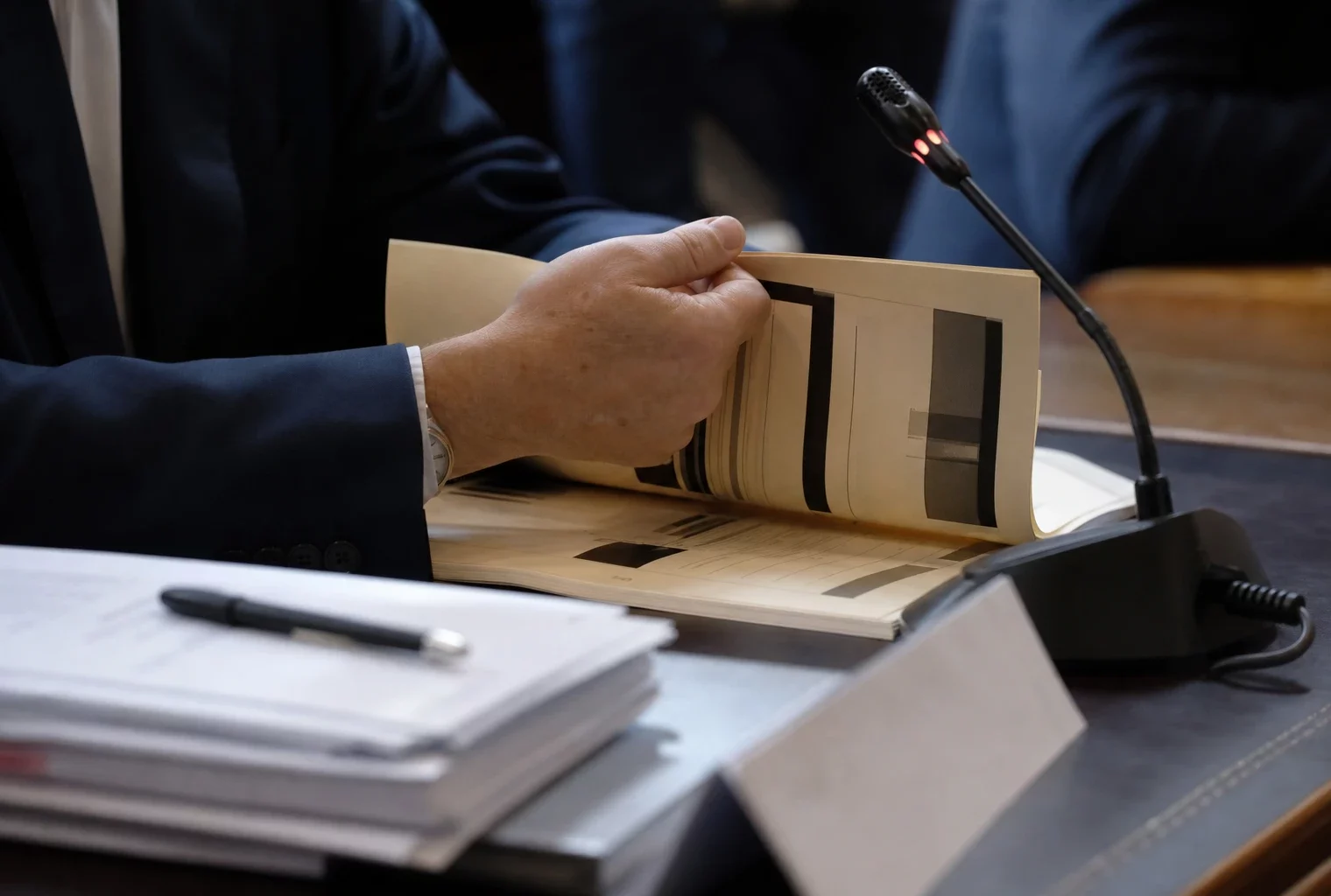 Main anonyme tournant un dossier de nomination devant un micro de commission parlementaire, en lumière naturelle.