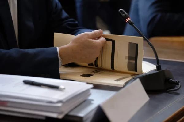 Main anonyme tournant un dossier de nomination devant un micro de commission parlementaire, en lumière naturelle.