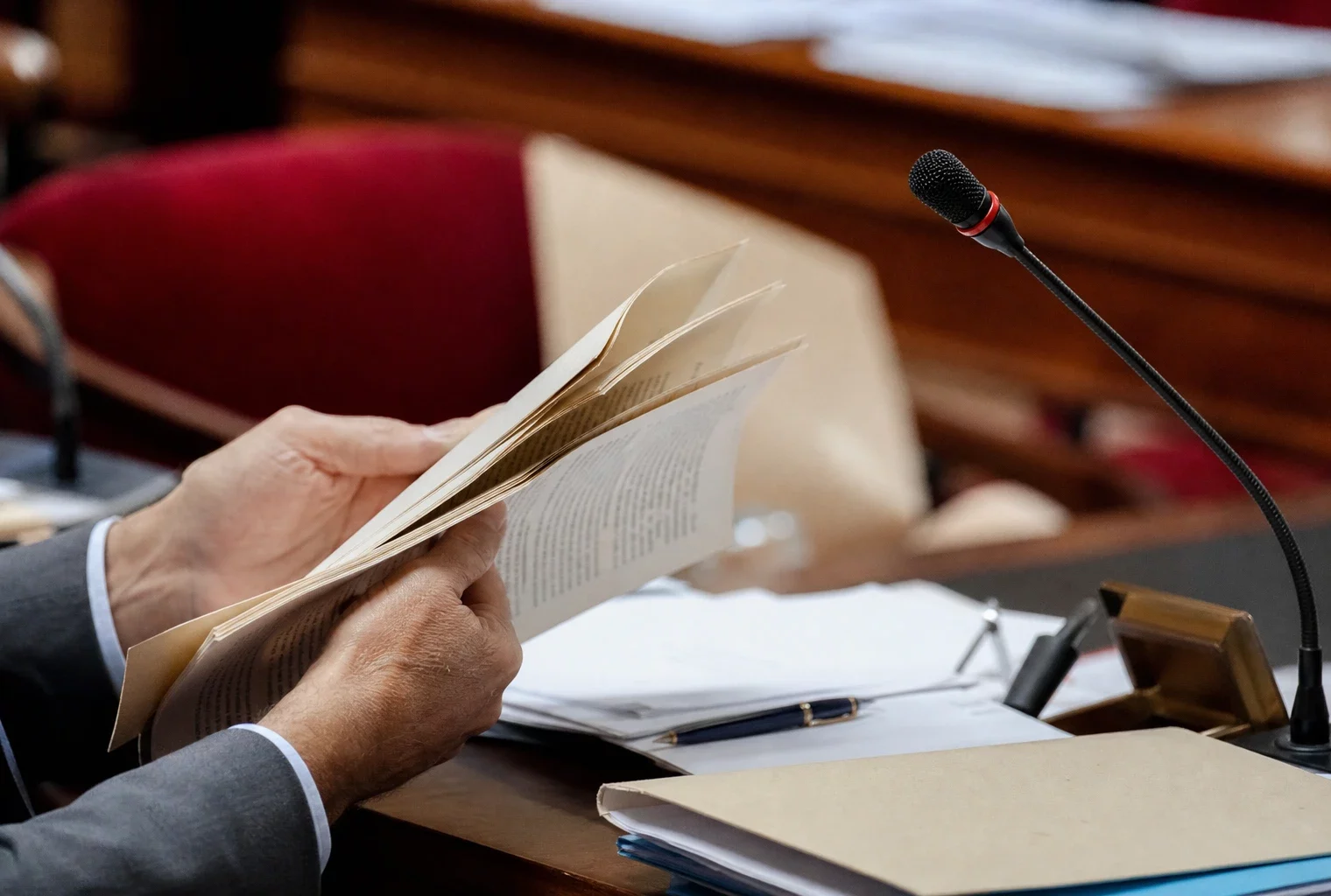 Mains anonymes tournant un dossier près d’un micro de commission dans une salle parlementaire française.