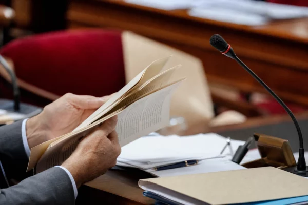 Mains anonymes tournant un dossier près d’un micro de commission dans une salle parlementaire française.