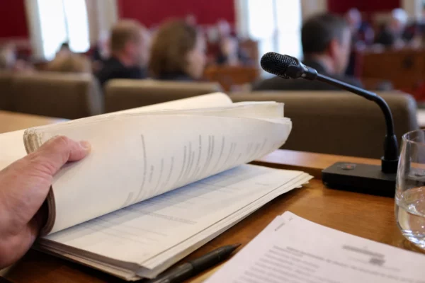 Dossier ministériel et micro de commission vus de près, avec une main feuilletant des documents flous dans une salle institutionnelle.
