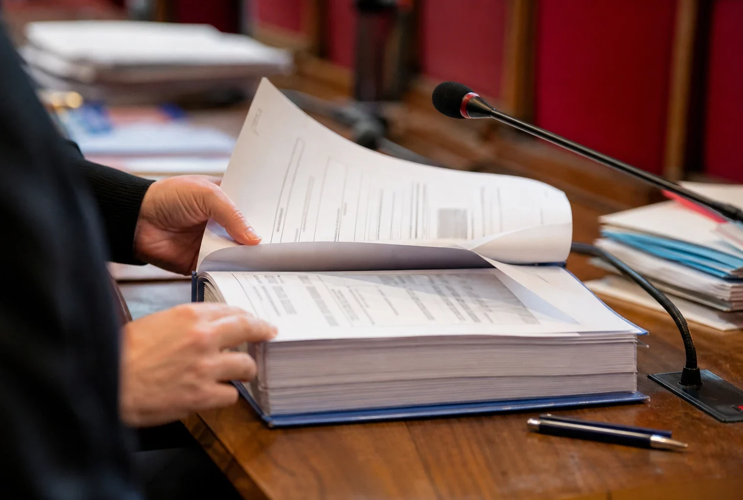 Dossier judiciaire et micro de commission dans une salle d’audition parlementaire, avec des mains anonymes et des papiers flous.