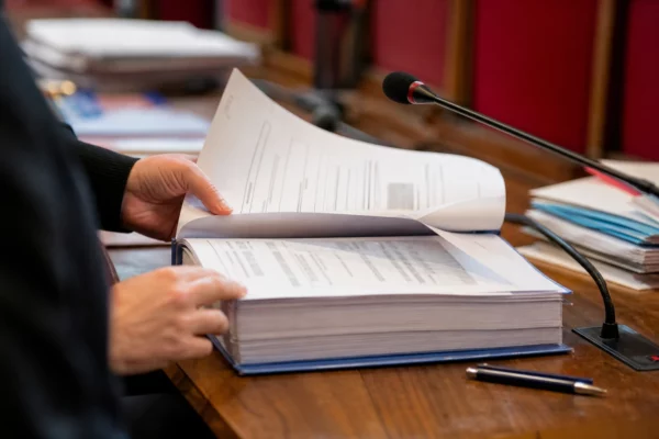 Dossier judiciaire et micro de commission dans une salle d’audition parlementaire, avec des mains anonymes et des papiers flous.