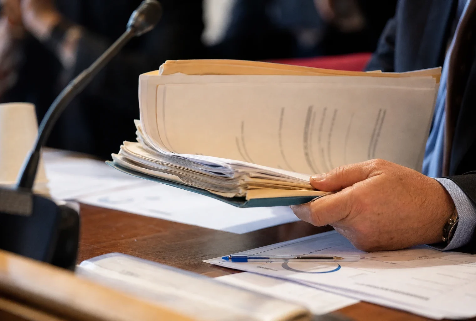 Dossier d’audition manipulé sur une table institutionnelle avec micro de commission et documents flous