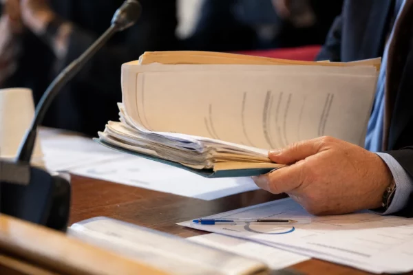 Dossier d’audition manipulé sur une table institutionnelle avec micro de commission et documents flous