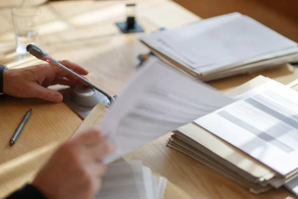 Main anonyme feuilletant un dossier de commission avec micro de séance et documents flous, en lumière naturelle.