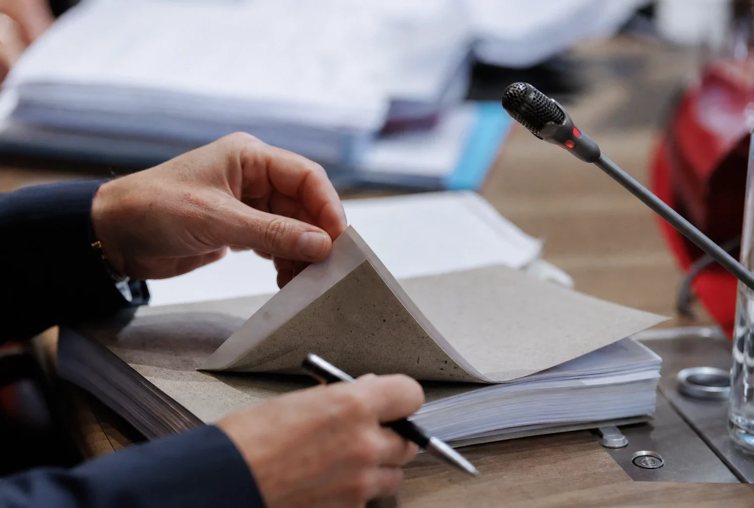 Mains anonymes feuilletant un dossier devant un micro de commission dans une salle institutionnelle française.