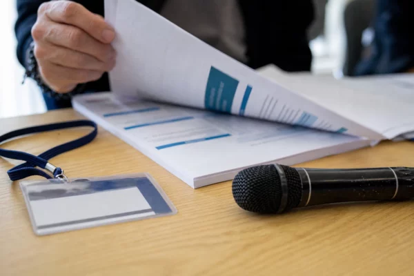 Gros plan d’un dossier institutionnel et d’un micro de presse sur une table, avec une main en train de feuilleter un document.