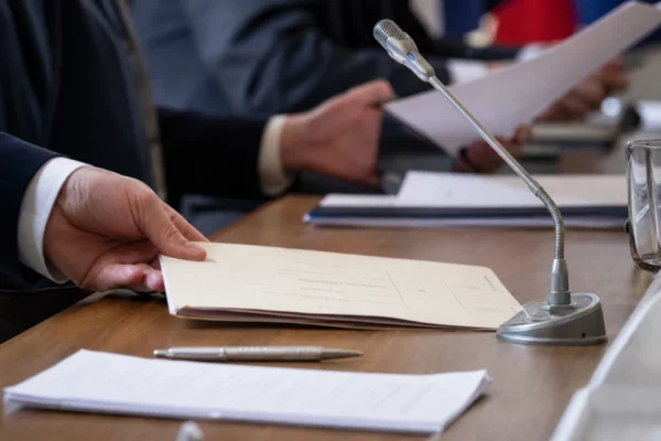 Dossier de nomination examiné sur une table de commission, avec micro et mains anonymes dans une lumière de fenêtre claire.