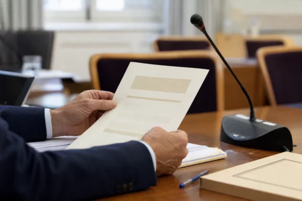 Dossier de nomination tenu par des mains anonymes dans une salle de commission avec micro et documents flous