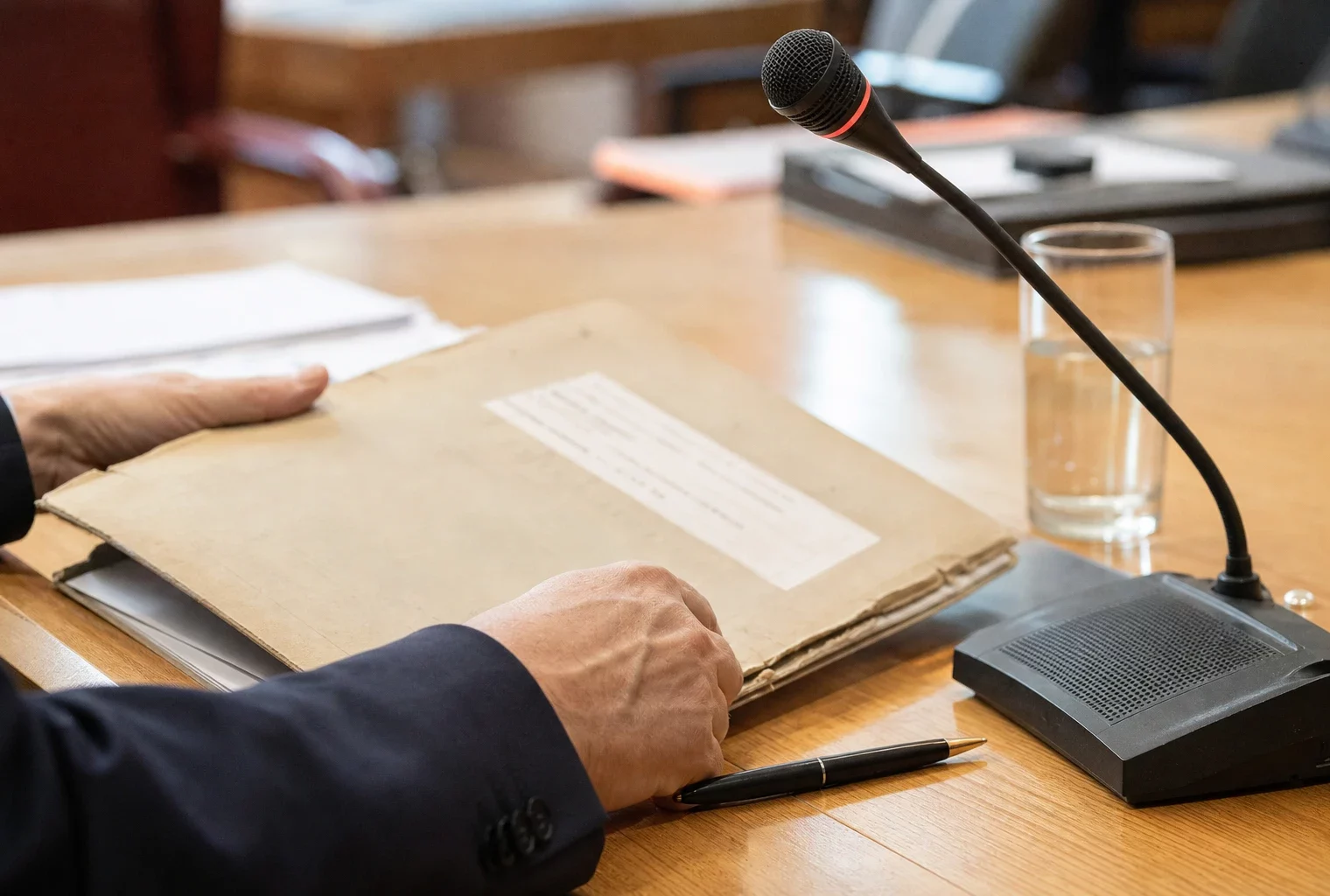 Main anonyme préparant un dossier de nomination près d’un micro de commission dans une salle institutionnelle lumineuse.