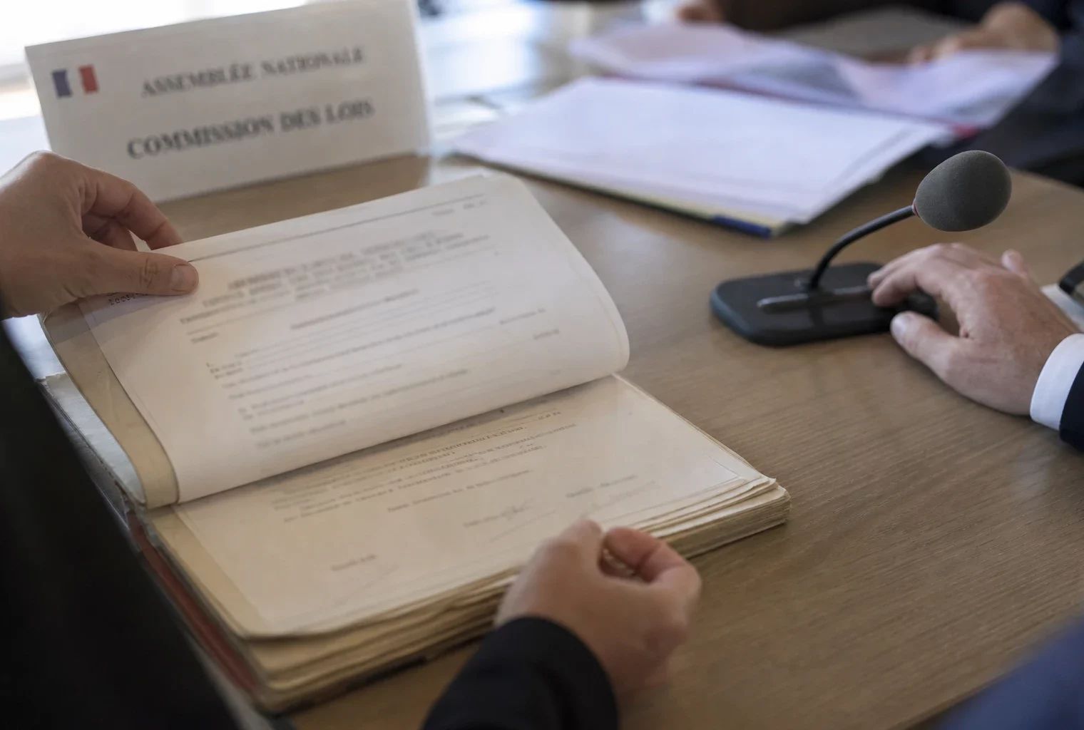 Dossier de nomination examiné par des mains anonymes devant un micro de commission, en lumière naturelle claire.