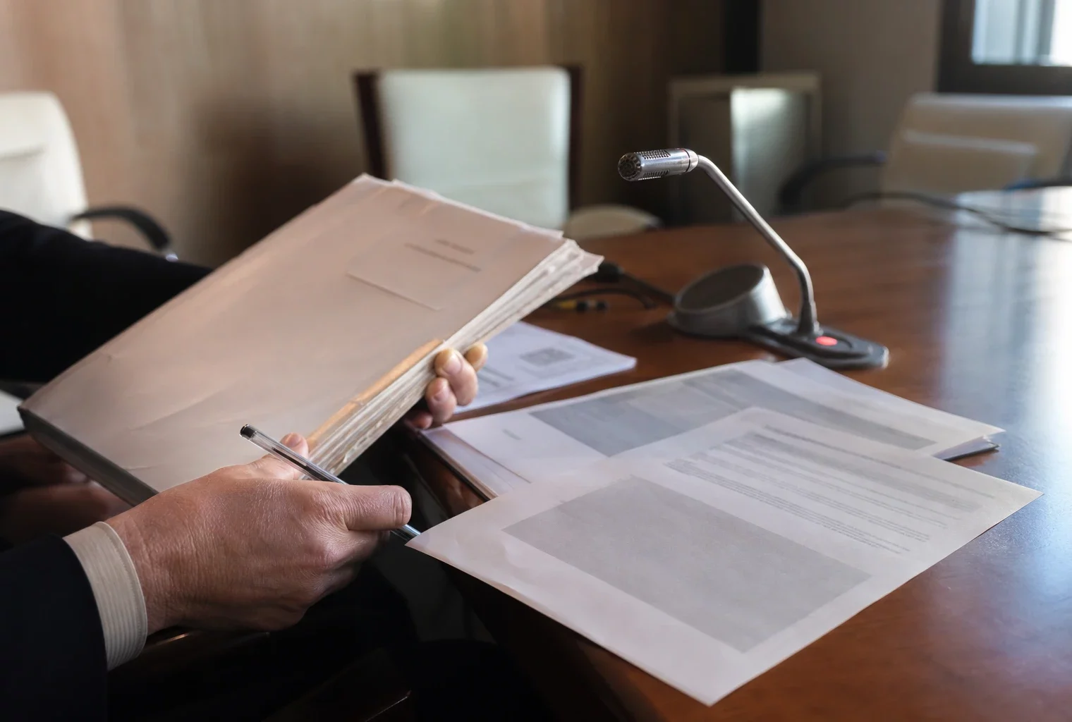 Mains anonymes sur un dossier de nomination près d’un micro de commission, dans une salle institutionnelle lumineuse.