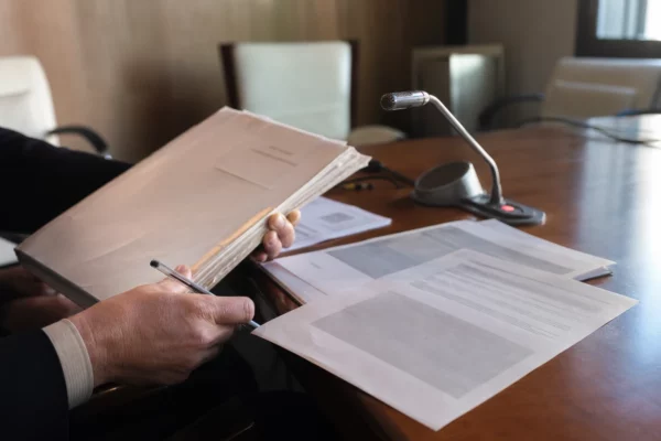 Mains anonymes sur un dossier de nomination près d’un micro de commission, dans une salle institutionnelle lumineuse.
