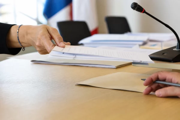 Main anonyme feuilletant un dossier de parrainage près d’un micro de commission dans une salle civique lumineuse.