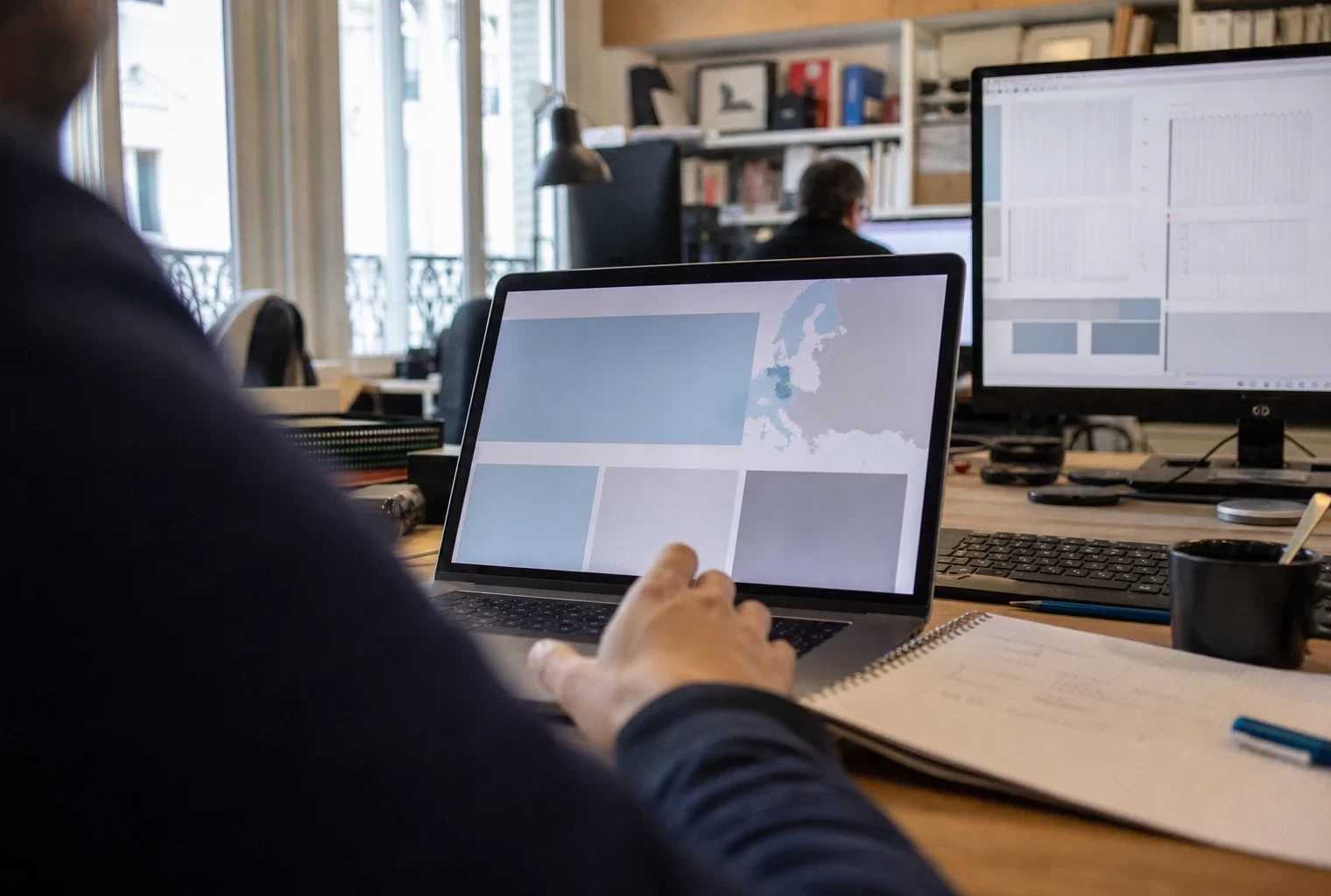 Vue d’un bureau de rédaction avec un écran d’ordinateur affichant une interface européenne abstraite et une main floue au premier plan