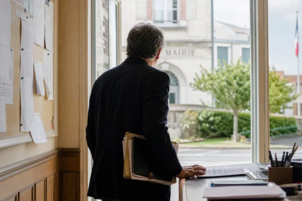 Élu local de dos dans une mairie, carnet et dossier à la main, ambiance calme de préparation budgétaire.