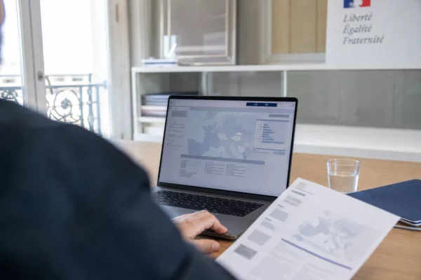 Vue par-dessus l’épaule d’un bureau parisien avec ordinateur affichant une interface européenne abstraite et un document flou.