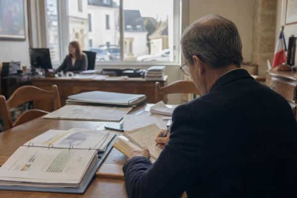 Un élu local de dos prend des notes dans une mairie, avec dossiers ouverts et lumière naturelle.