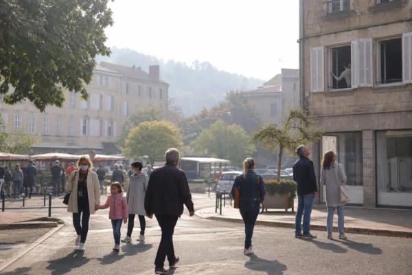 Des habitants traversent une place de ville moyenne sous une légère fumée de feu de forêt, près de la mairie.