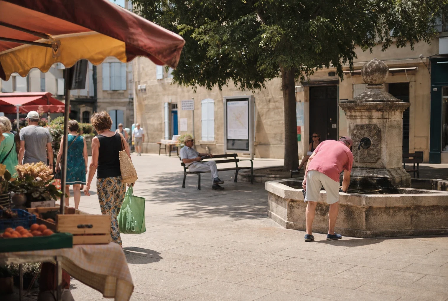 Place de ville moyenne en pleine journée, passants anonymes à l’ombre et ambiance calme sous la chaleur