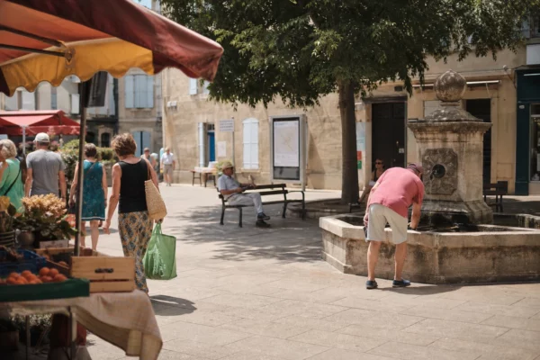 Place de ville moyenne en pleine journée, passants anonymes à l’ombre et ambiance calme sous la chaleur