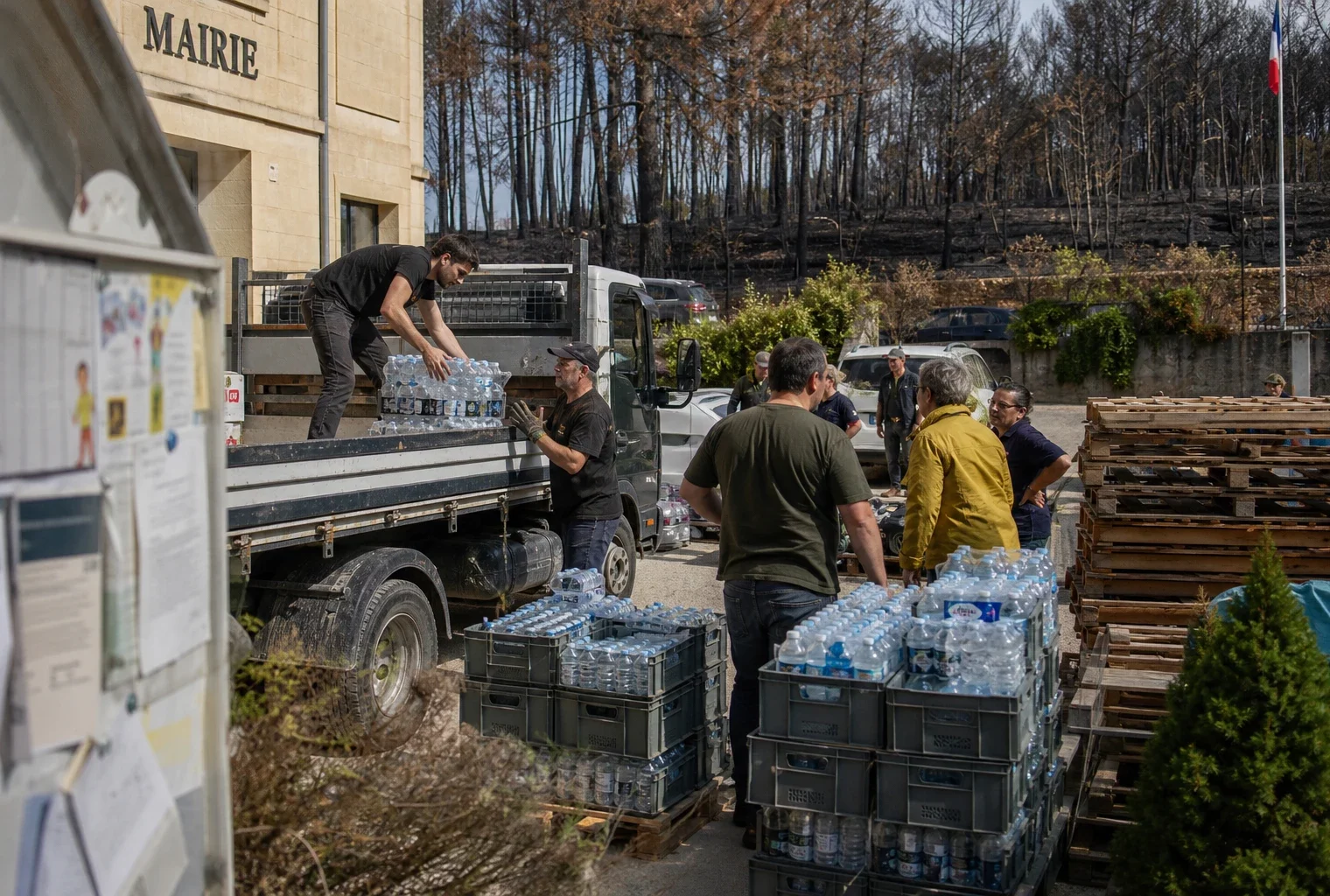 Des habitants et bénévoles déchargent des bouteilles d’eau près d’une mairie en Gironde, avec une forêt marquée par l’incendie au loin.