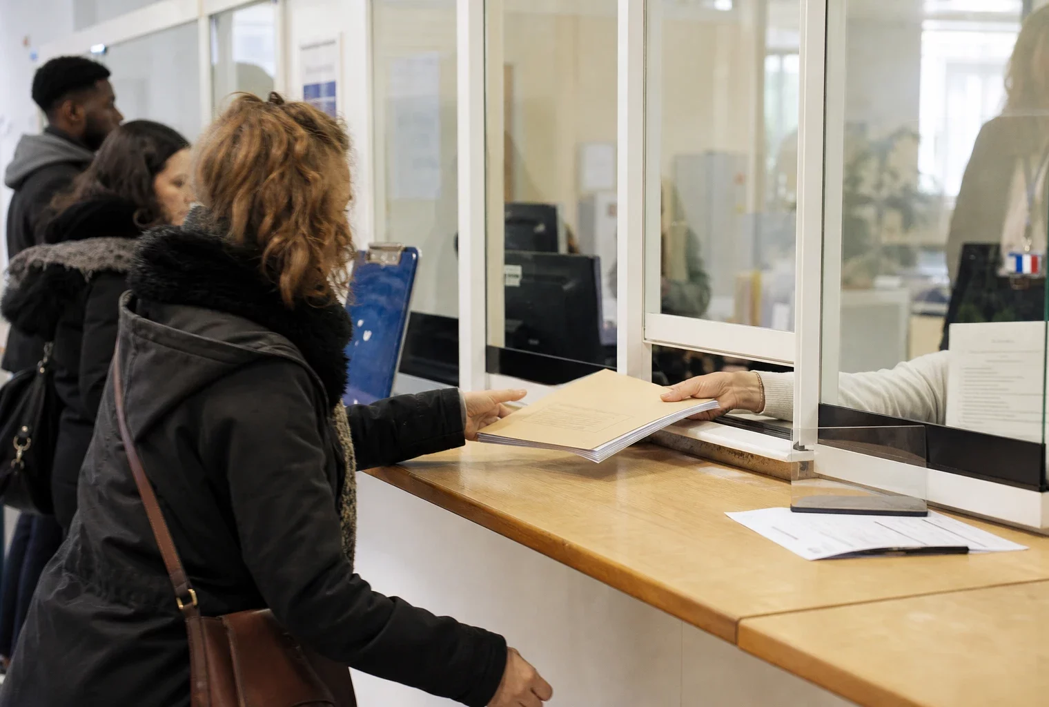 Guichet d’accueil administratif français avec dossier, main et usagers anonymes dans une lumière claire.