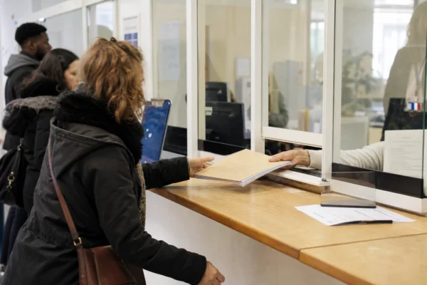 Guichet d’accueil administratif français avec dossier, main et usagers anonymes dans une lumière claire.