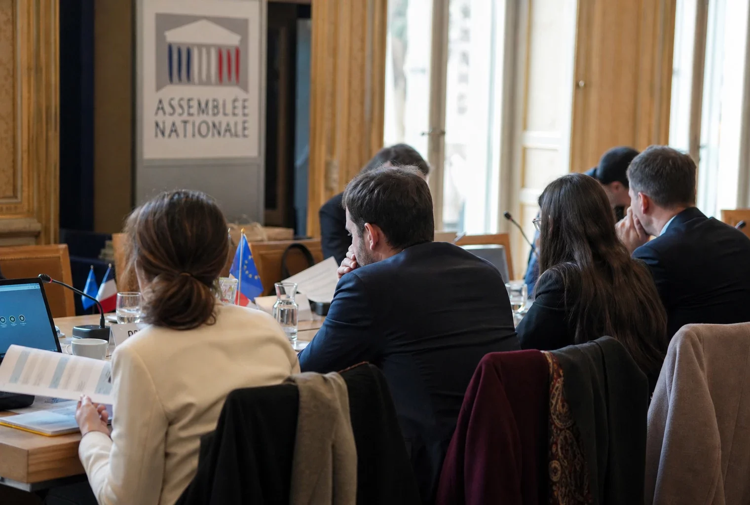 Réunion institutionnelle sobre avec dossiers et micros, silhouettes de dos autour d’une table européenne