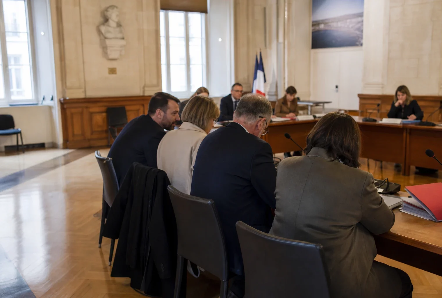 Salle municipale lumineuse avec micros, dossiers flous et une chaise vide lors d’une réunion locale