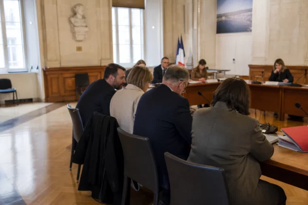Salle municipale lumineuse avec micros, dossiers flous et une chaise vide lors d’une réunion locale