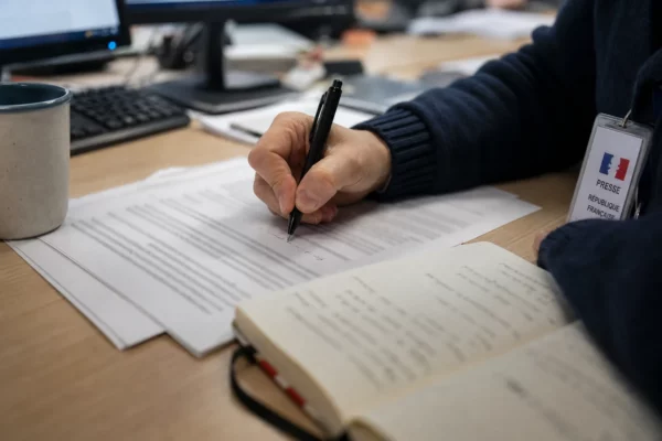 Main de journaliste annotant une page imprimée floue dans une rédaction française, avec carnet et lumière de fenêtre.