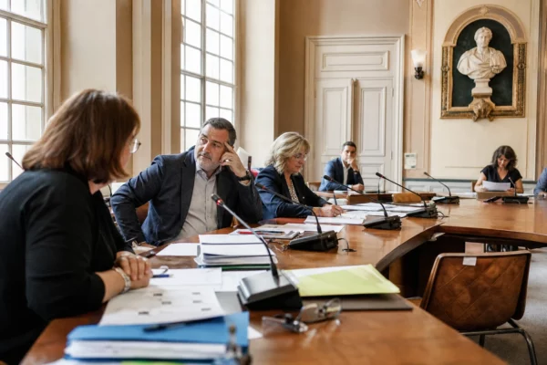Salle municipale lumineuse avec micros, dossiers flous et chaise vide lors d’un débat local sur la loi agricole