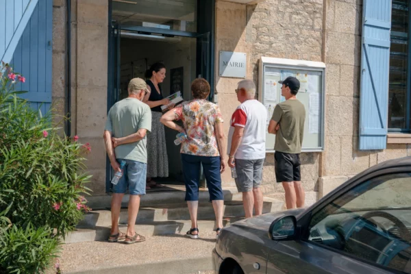 Des habitants discutent devant une mairie française par forte chaleur, dans une scène de vie citoyenne ordinaire.