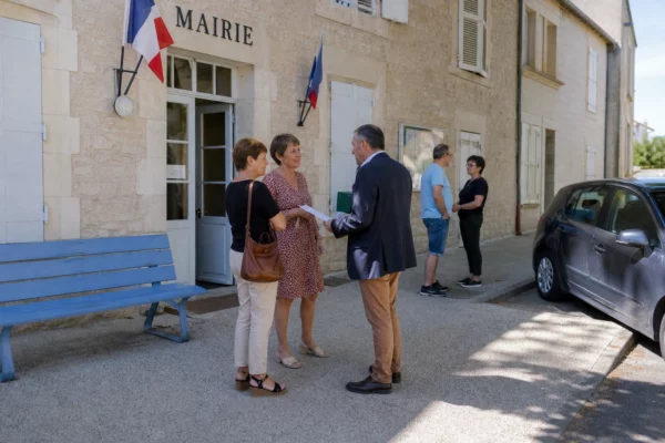 Des habitants discutent devant la mairie d’une petite ville, sous une lumière naturelle claire et quotidienne.